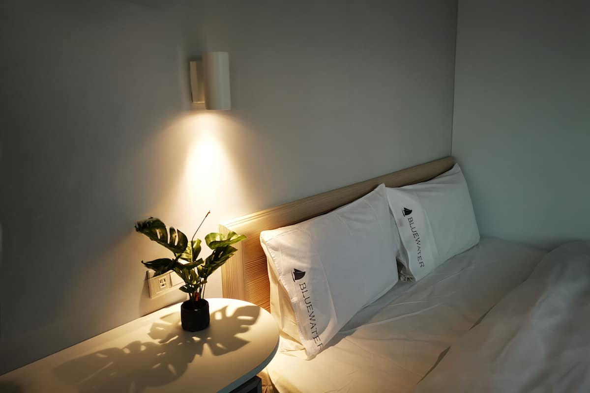 Habitación de hotel con cama, almohadas blancas, mesita de noche con una planta y una lámpara de pared.