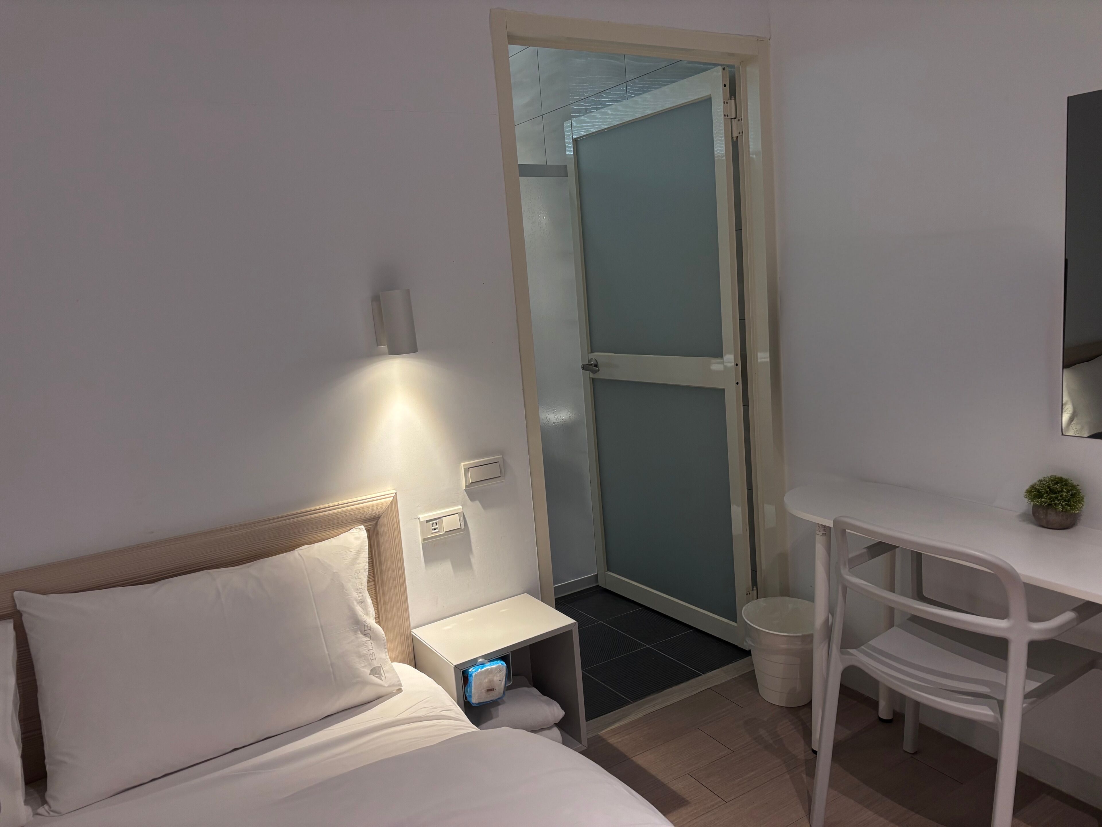 Habitación de hotel con cama, un pequeño escritorio, una silla, una mesita de noche, un cubo de basura y una puerta que da a un baño.