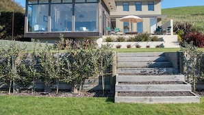 A modern house with a large glass conservatory, a patio with outdoor furniture, and a well-maintained garden with raised beds. - Kotare Point Paradise - Akaroa Holiday Home (Akaroa)