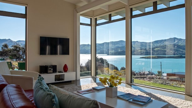 A modern living room with a large window offering a view of a lake and mountains. - Kotare Point Paradise - Akaroa Holiday Home (Akaroa)