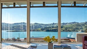 A room with large windows offering a view of a lake and mountains. - Kotare Point Paradise - Akaroa Holiday Home (Akaroa)
