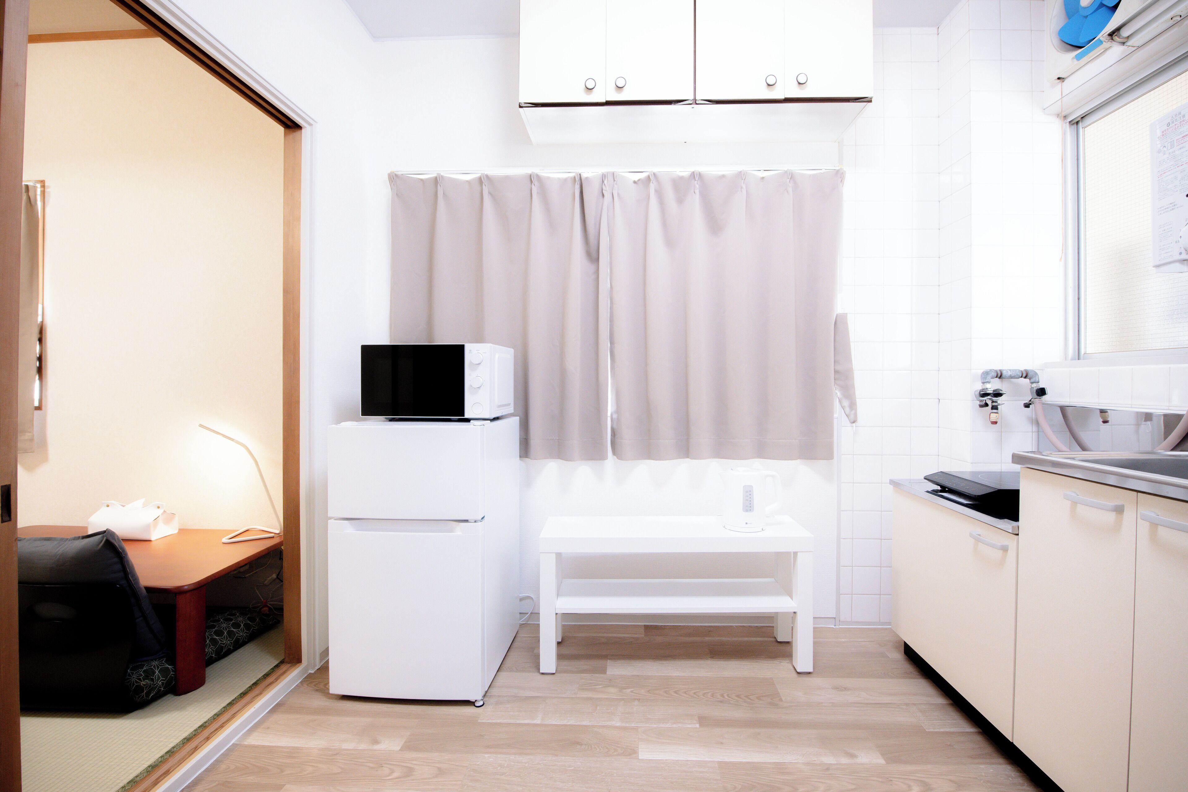 Cocina compacta con microondas, refrigerador pequeño, banco blanco y ventana con cortinas.