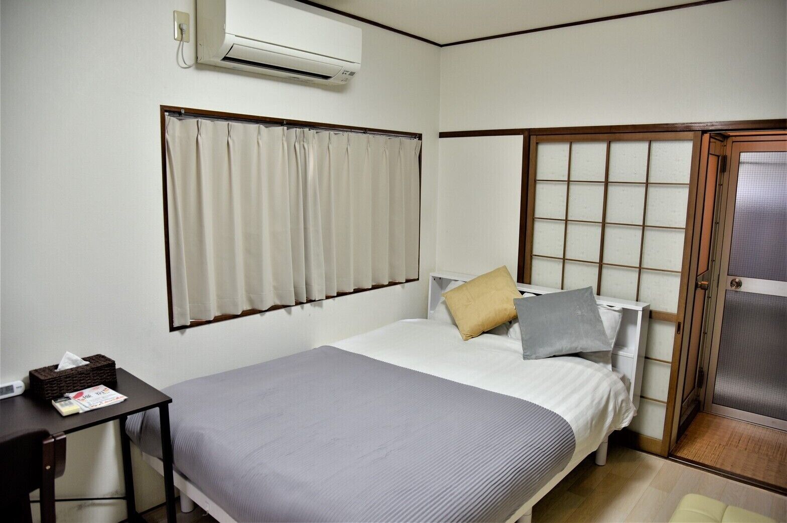 A bedroom with a bed, a bedside table, an air conditioning unit, a window with curtains, and a sliding door.