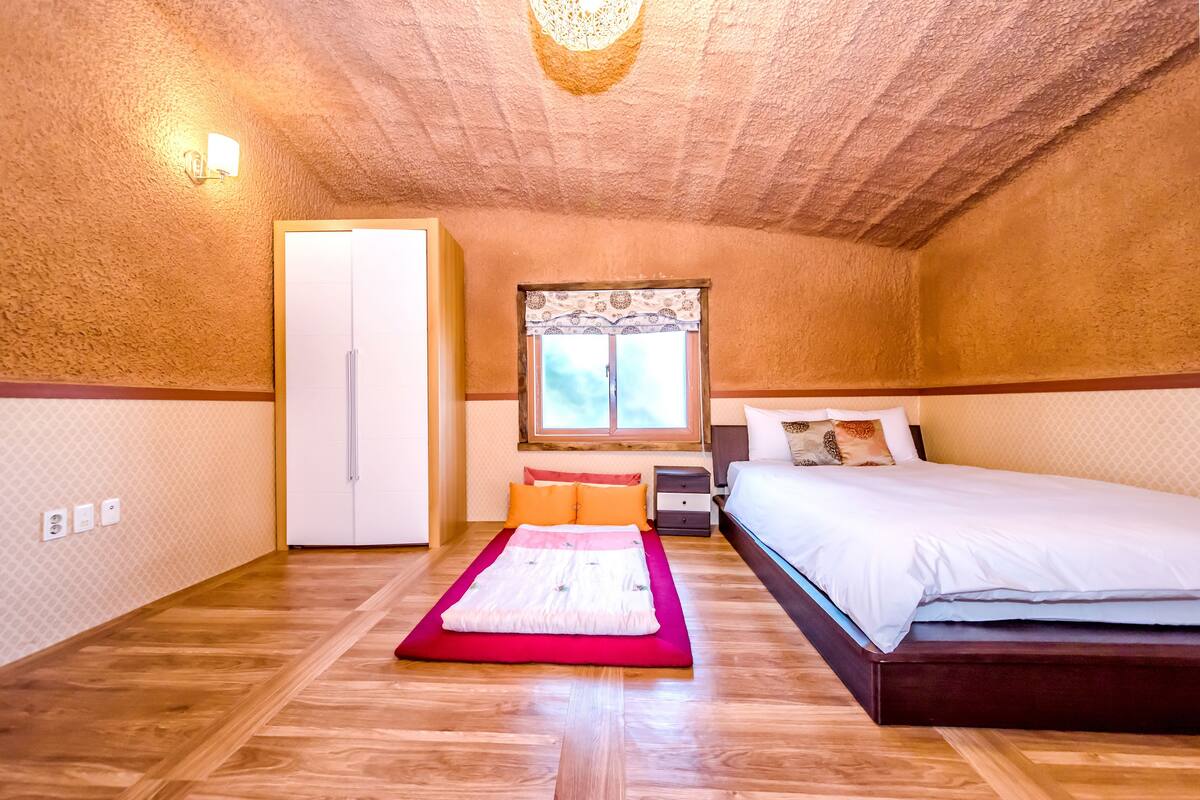 A bedroom with a wooden floor, a bed with white bedding, a nightstand, a dresser, a window with patterned curtains, and a ceiling light fixture.