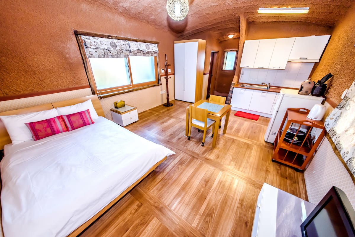 A compact room with a bed, a small table, a chair, a kitchenette, and a wardrobe.