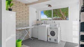 Een praktische wasruimte met een wasmachine, droger en een wasbak. Een raam zorgt voor natuurlijk licht, en een groene plant geeft een vleugje natuur.