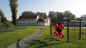 A grassy area with a playground structure and a pathway leading to a building with a red roof. - Park Atlantis (De Haan)