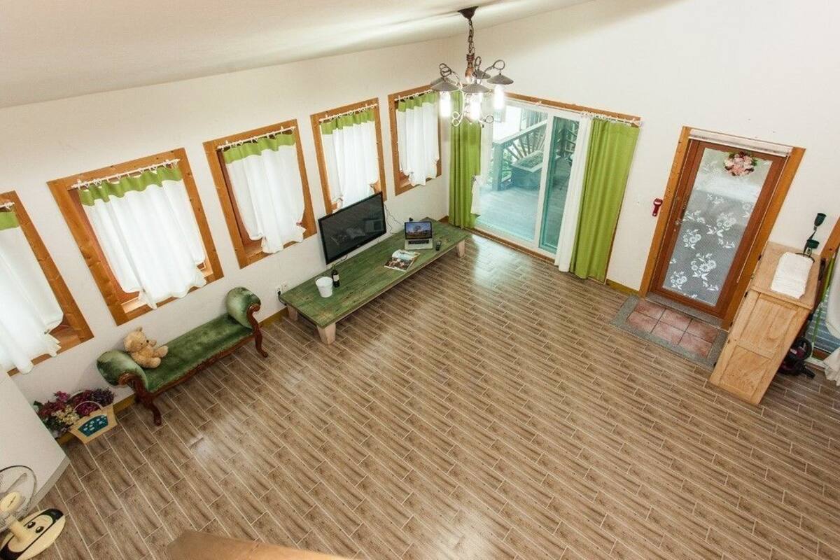 Ein geräumiges Wohnzimmer mit Holzboden, einem grünen Sofa, einem Holztisch, einem Fernseher und einer Glastür, die auf einen Balkon führt.