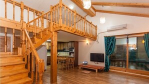 A wooden staircase leading to an upper level, a dining area with chairs, a television, and a large window with curtains. - Jirisan Shiri Pension (Gurye)