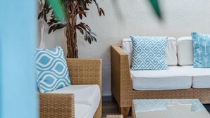 Un sofá de mimbre con cojines blancos y almohadas estampadas, una planta en maceta y un tapete con textura.