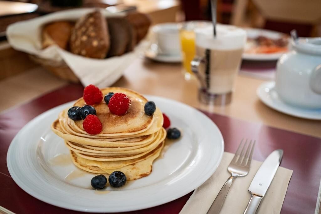 En hög med pannkakor på en vit tallrik, dekorerade med blåbär och hallon, tillsammans med en gaffel och en kniv som ligger på en servett.