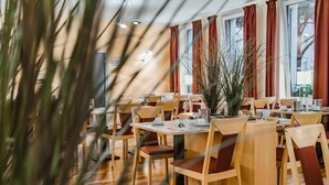 Un restaurante con mesas y sillas de madera, ventanas amplias con cortinas y plantas de interior.