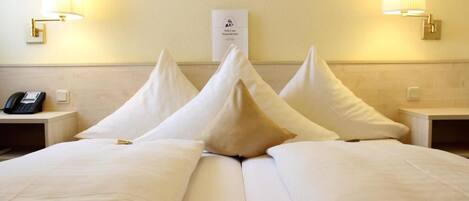 Habitación de hotel con una cama bien hecha, dos lámparas de noche, un teléfono en la mesita izquierda y un cartel en la pared.