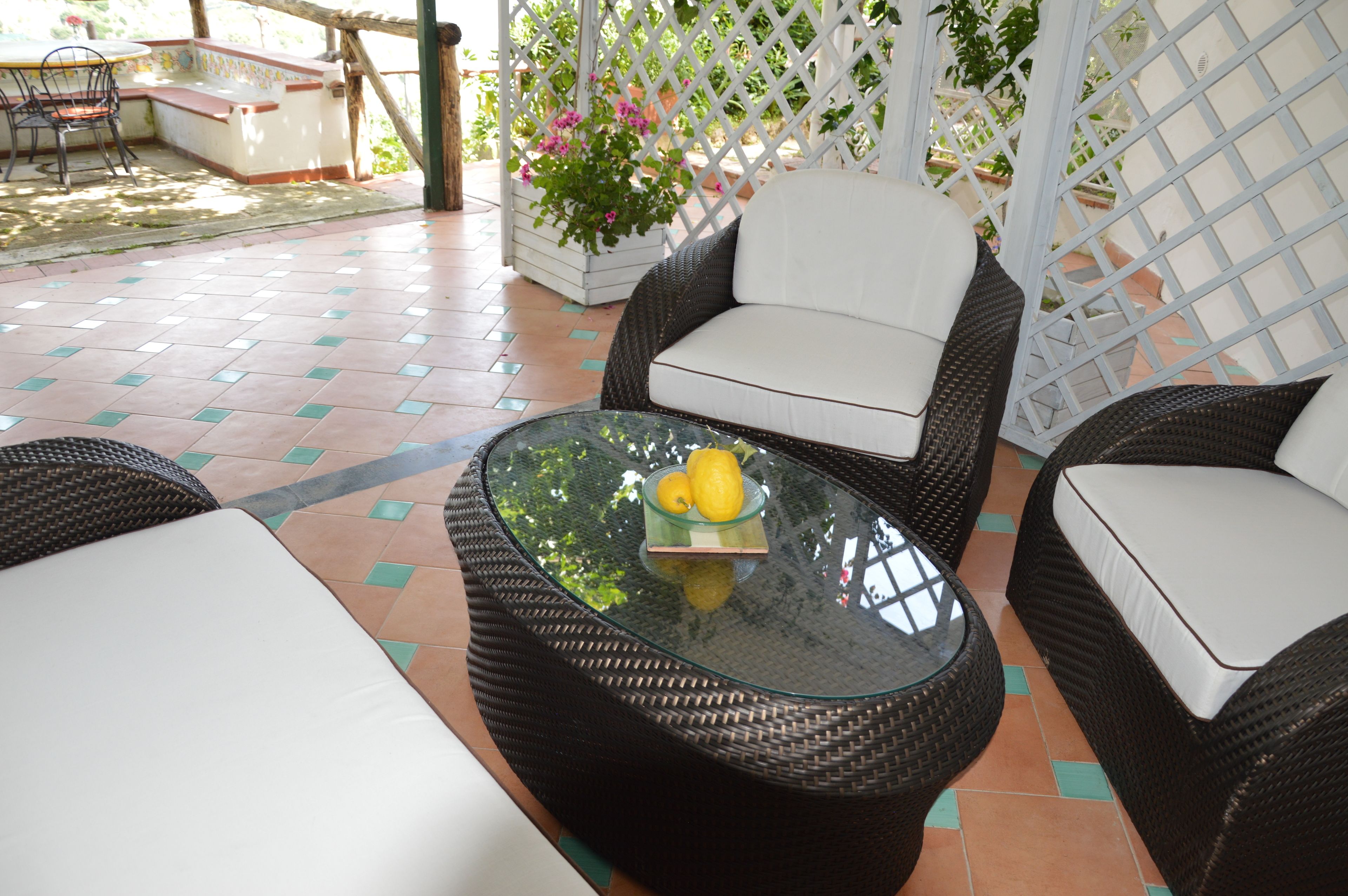 Un patio con mobiliario de mimbre, una mesa de vidrio con limones y una valla de celosía blanca.
