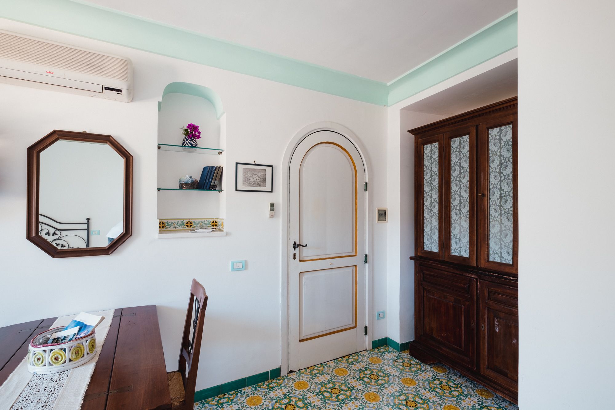 Une pièce comprenant une table à manger en bois, une chaise, une porte blanche ornée d’un cadre décoratif, un miroir, une étagère avec des livres et un vase, ainsi qu’un meuble avec des portes en verre.