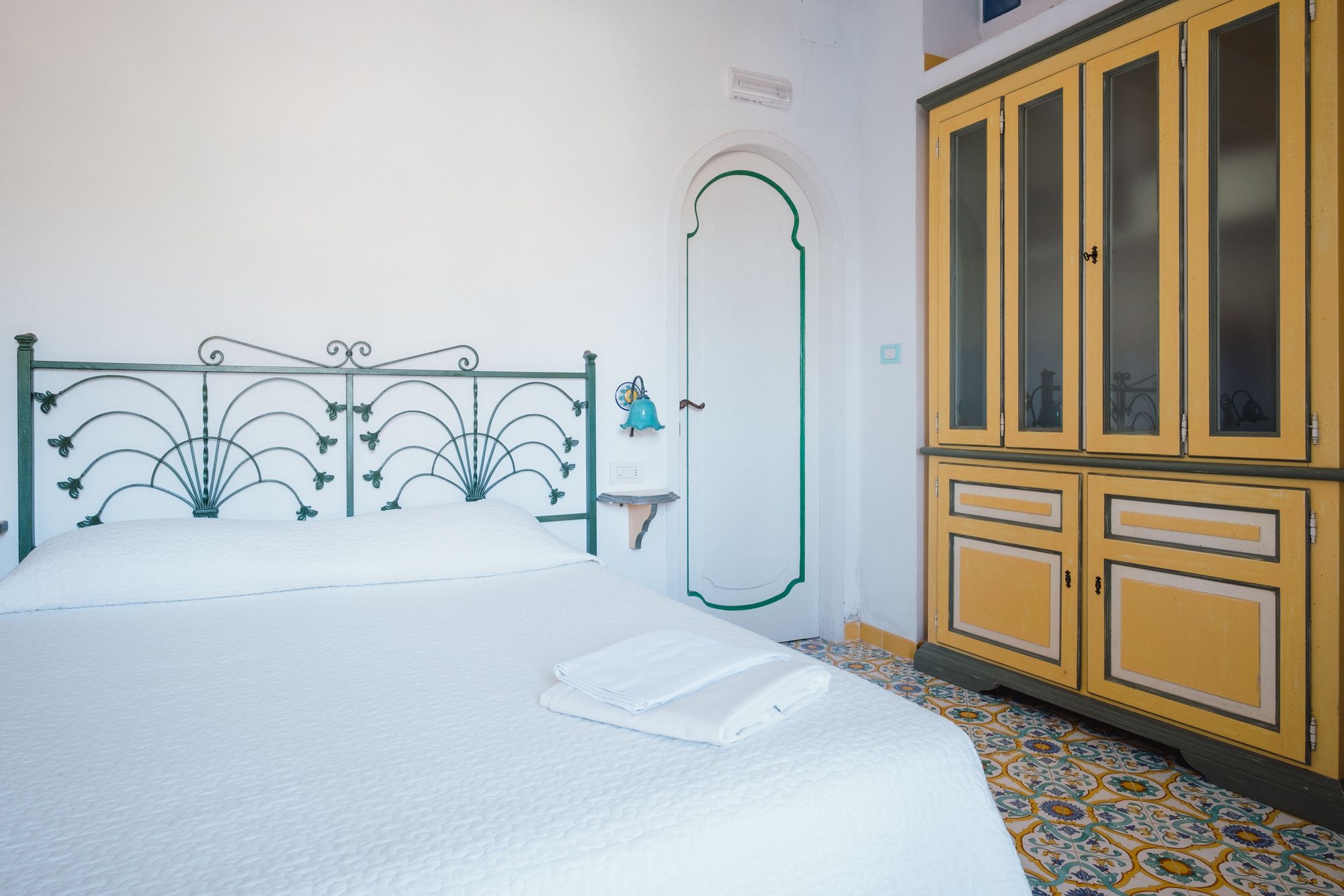 A neatly made bed with a white duvet, a teal wall-mounted bell, and a wooden cabinet with glass doors.