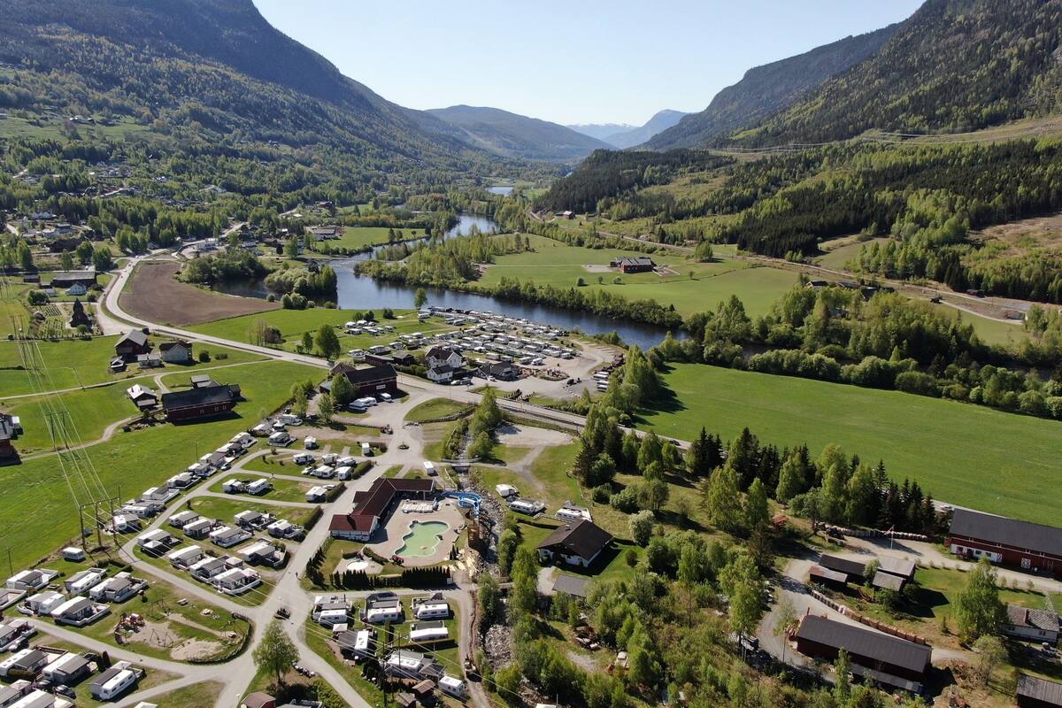 A campground with RVs and cabins, surrounded by green fields and a lake, with mountains in the background.