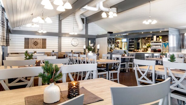 Un restaurant spacieux, doté de meubles blancs, d’un éclairage suspendu et d’une cuisine ouverte.