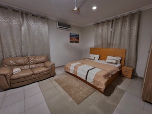 A bedroom with a bed, a leather sofa, a small wooden nightstand, and a wall-mounted air conditioner.