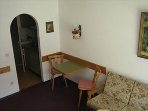 A small, cozy room with a wooden table and two chairs, a patterned sofa, and a small shelf with decorative items.