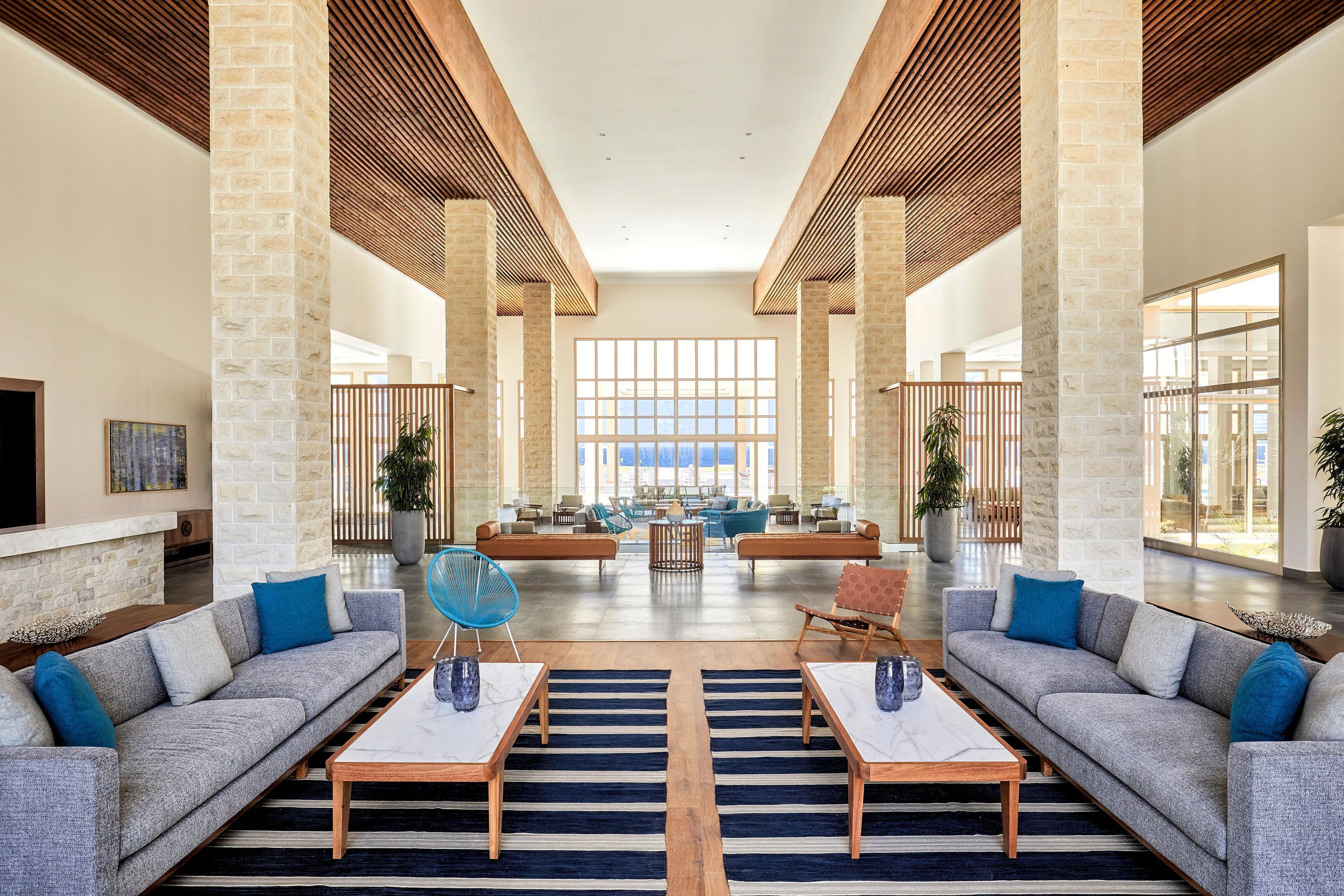 A spacious lobby with high ceilings, large windows, and a modern design featuring sofas, tables, and potted plants.