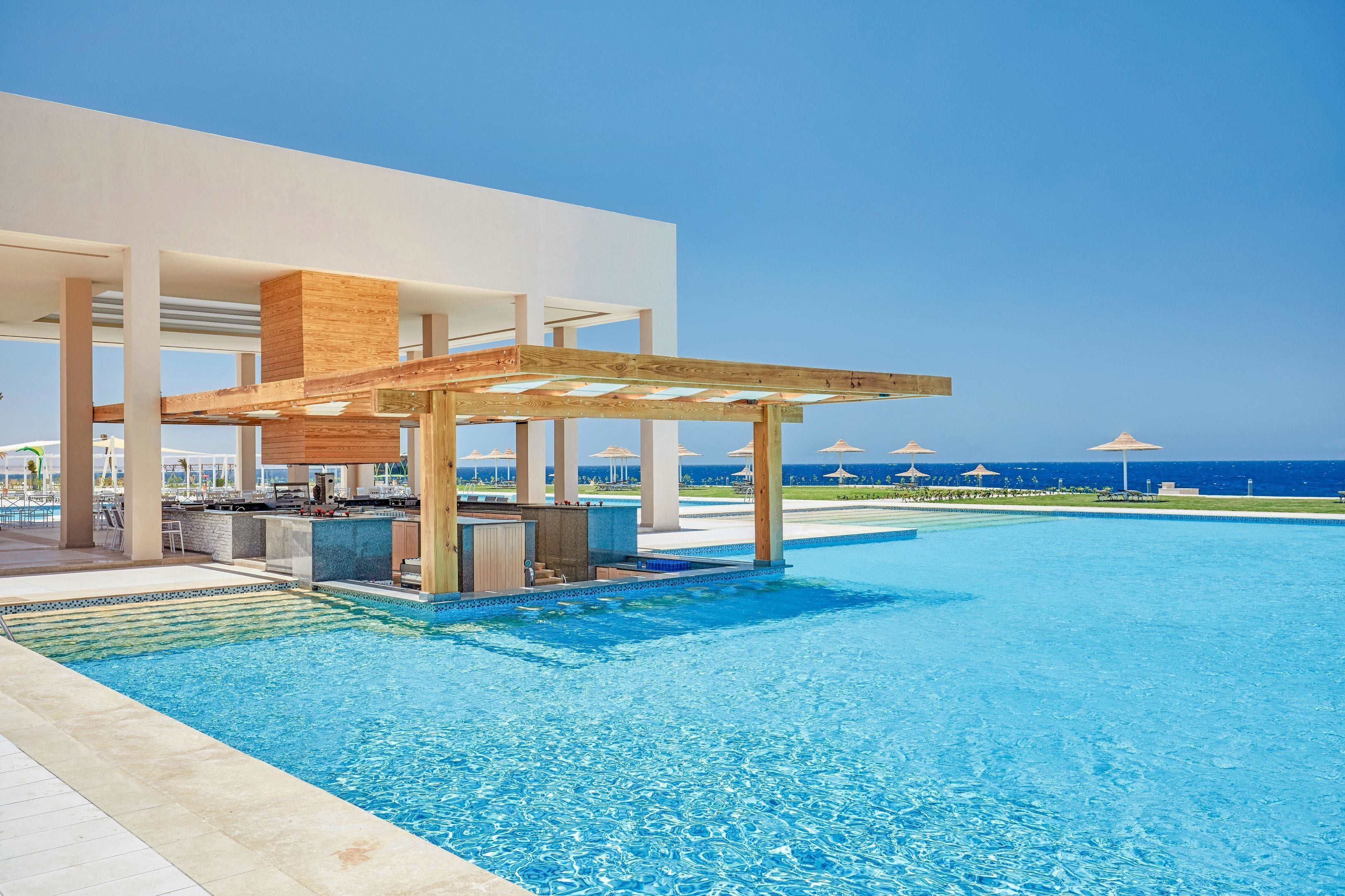 A modern pool area with a wooden pergola, lounge chairs, and a view of the ocean.