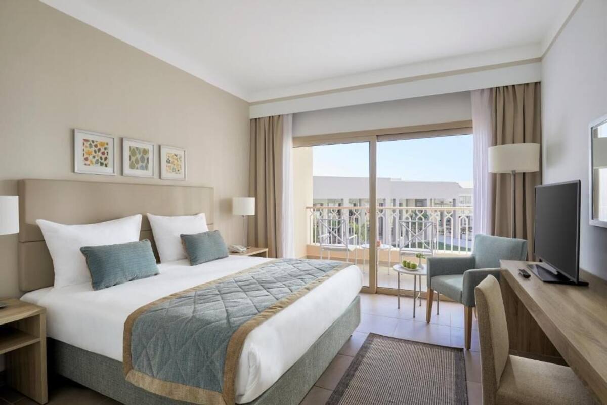 A hotel room with a bed, a desk, a chair, a TV, a balcony with a view, and artwork on the wall.
