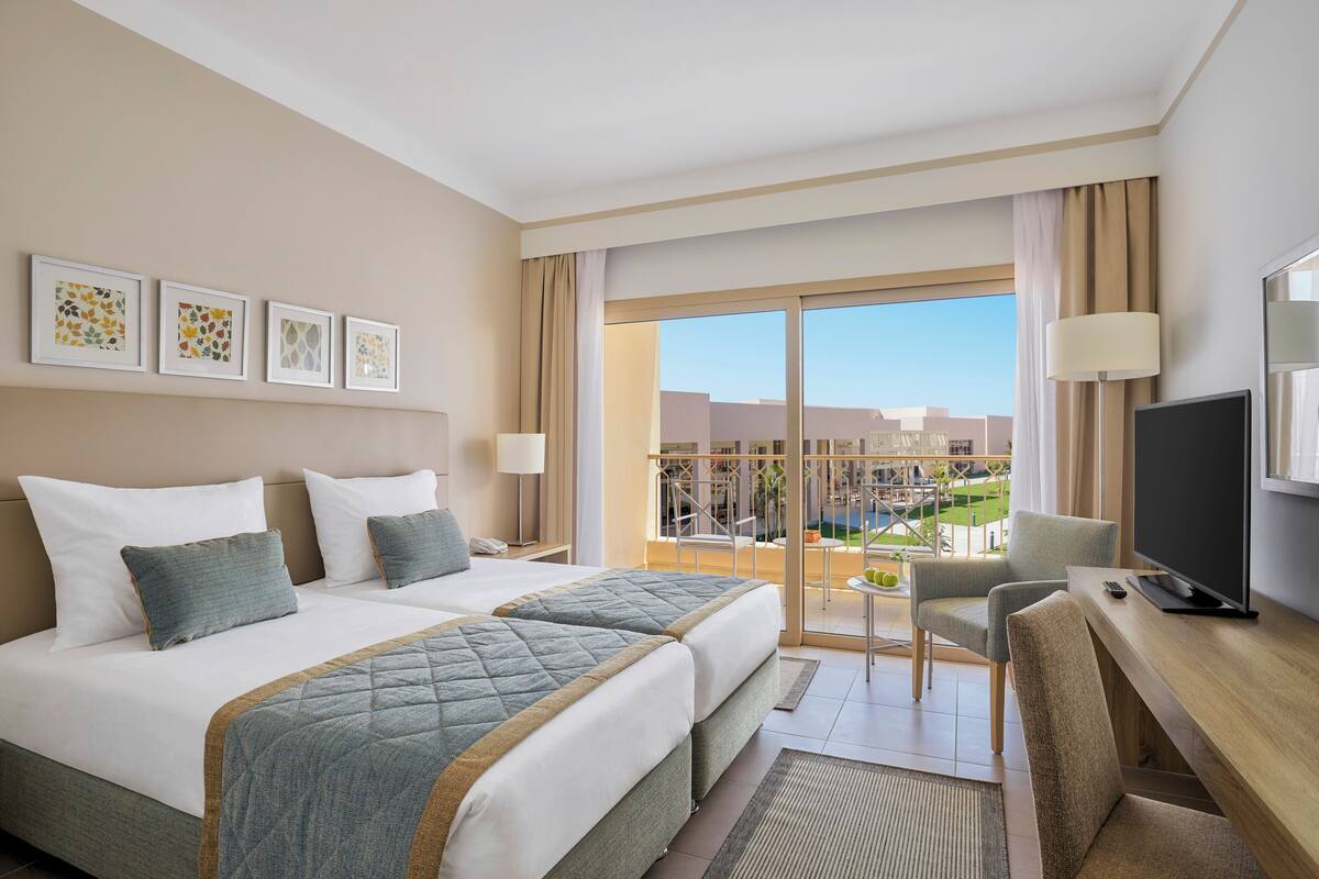 A hotel room with two beds, a desk, a TV, and a balcony with a view of buildings and greenery.