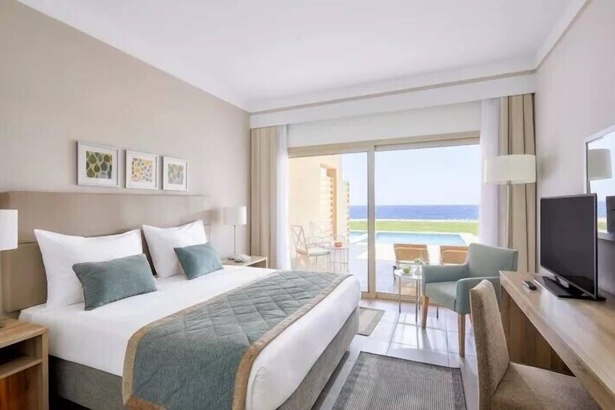 A hotel room with a bed, a desk, a TV, and a balcony with a view of the ocean.