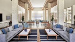 Een ruime lobby met een hoge plafond, grote ramen en een centraal gelegen zithoek met banken en tafels.