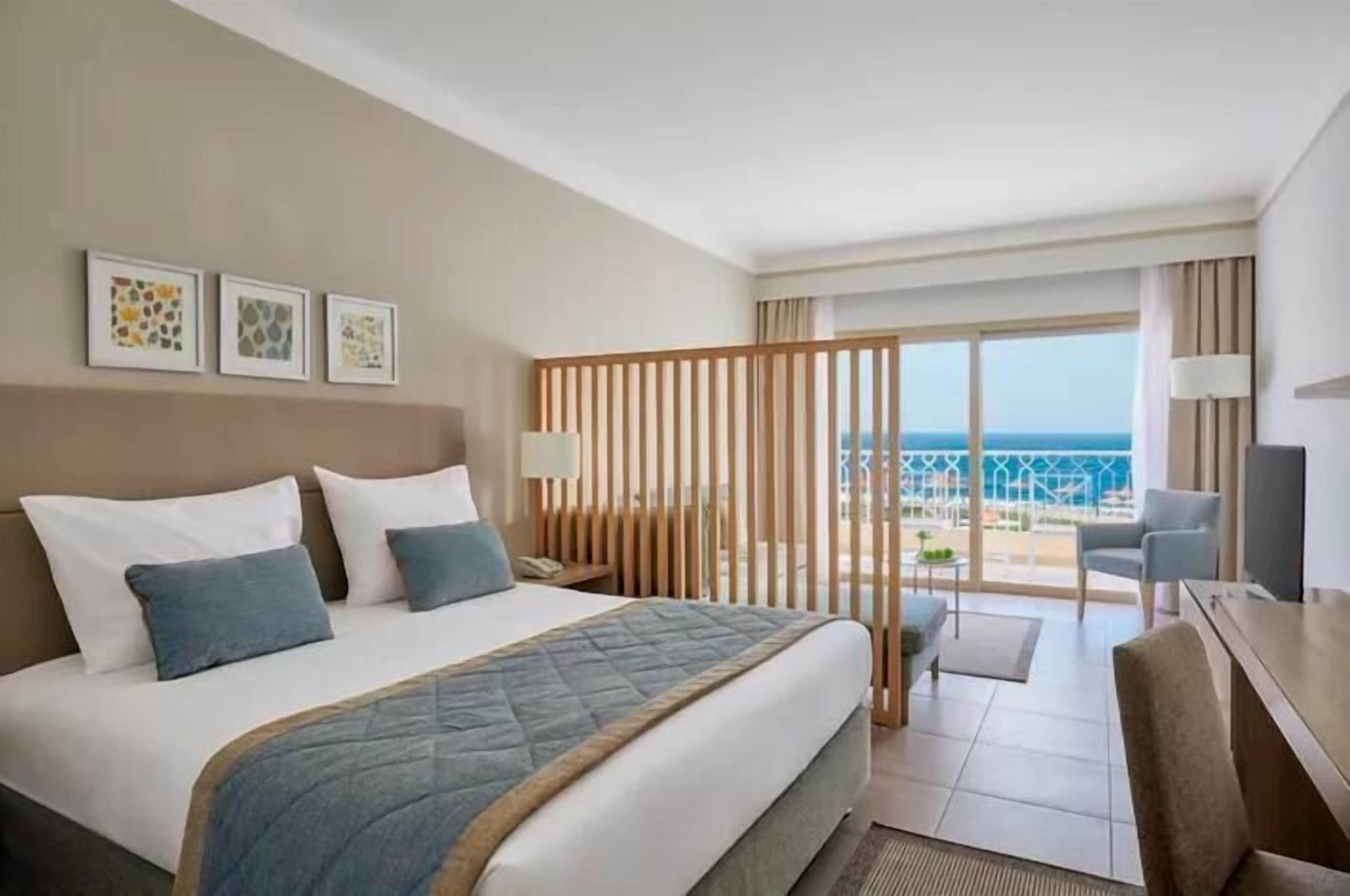 Habitación de hotel con cama, escritorio, silla, televisor y balcón con vistas al mar.