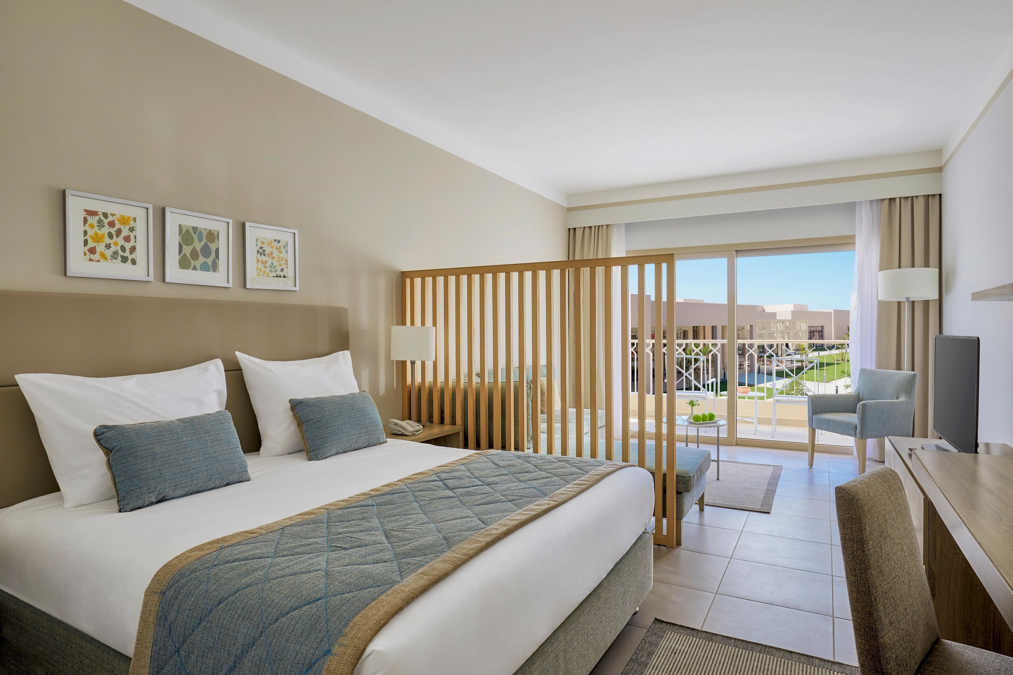 A hotel room with a large bed, a desk with a chair, a TV, and a balcony with a view.