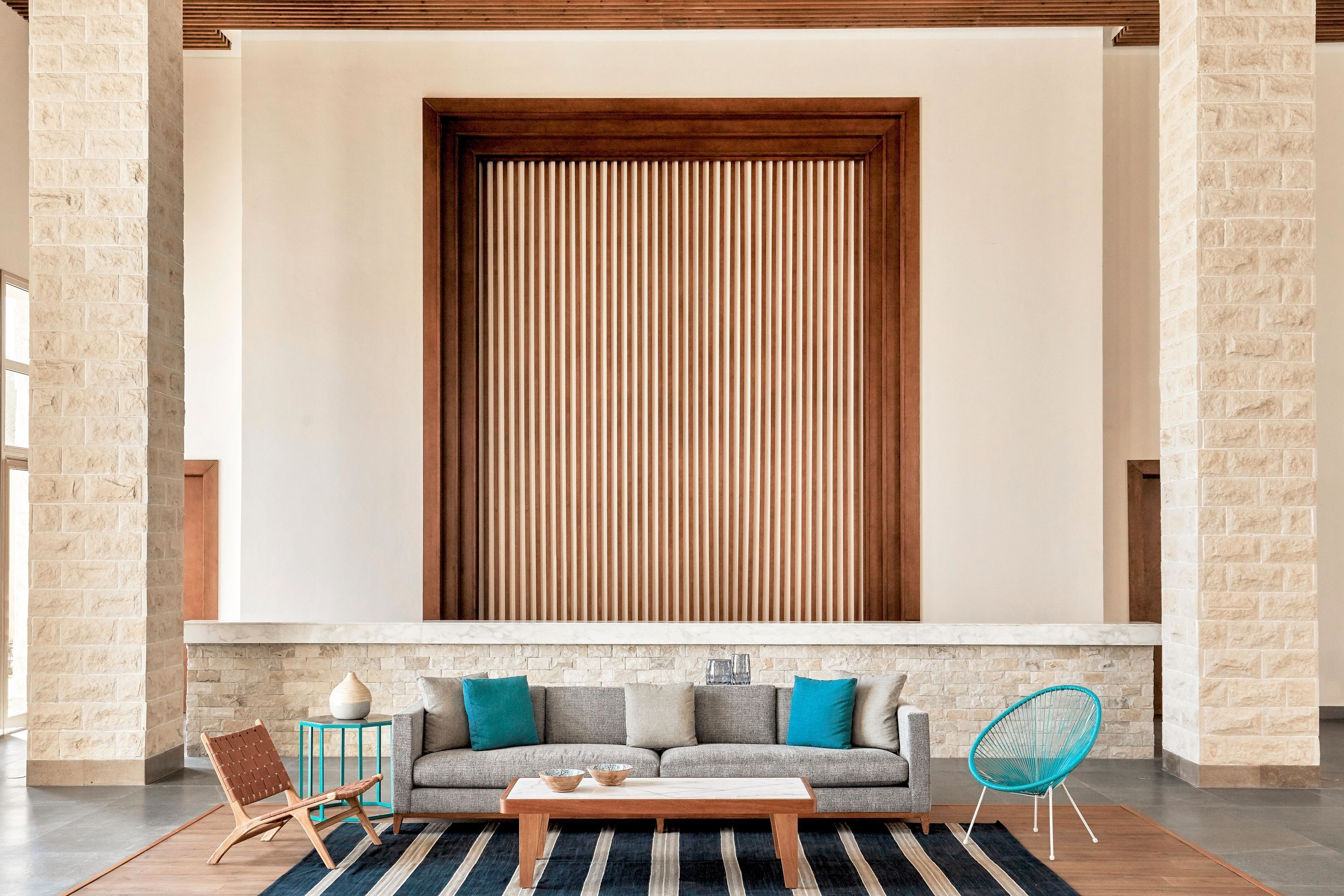 A modern living room with a stone wall, a wooden slatted panel, a grey sofa with blue cushions, a wooden coffee table, a blue chair, and a striped rug.