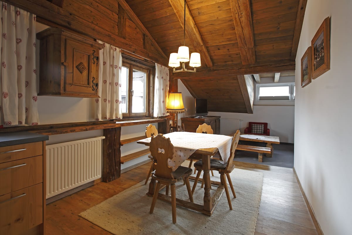 A rustic kitchen with wooden beams, a dining table with chairs, a radiator, and a window with curtains.