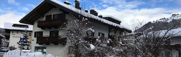 Ein traditionelles Chalet mit Holzbalkon, umgeben von schneebedeckten Bäumen und einer Bergkulisse.