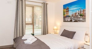 A hotel room with a bed, a painting of a bridge, a window with blinds, and a nightstand with a lamp.