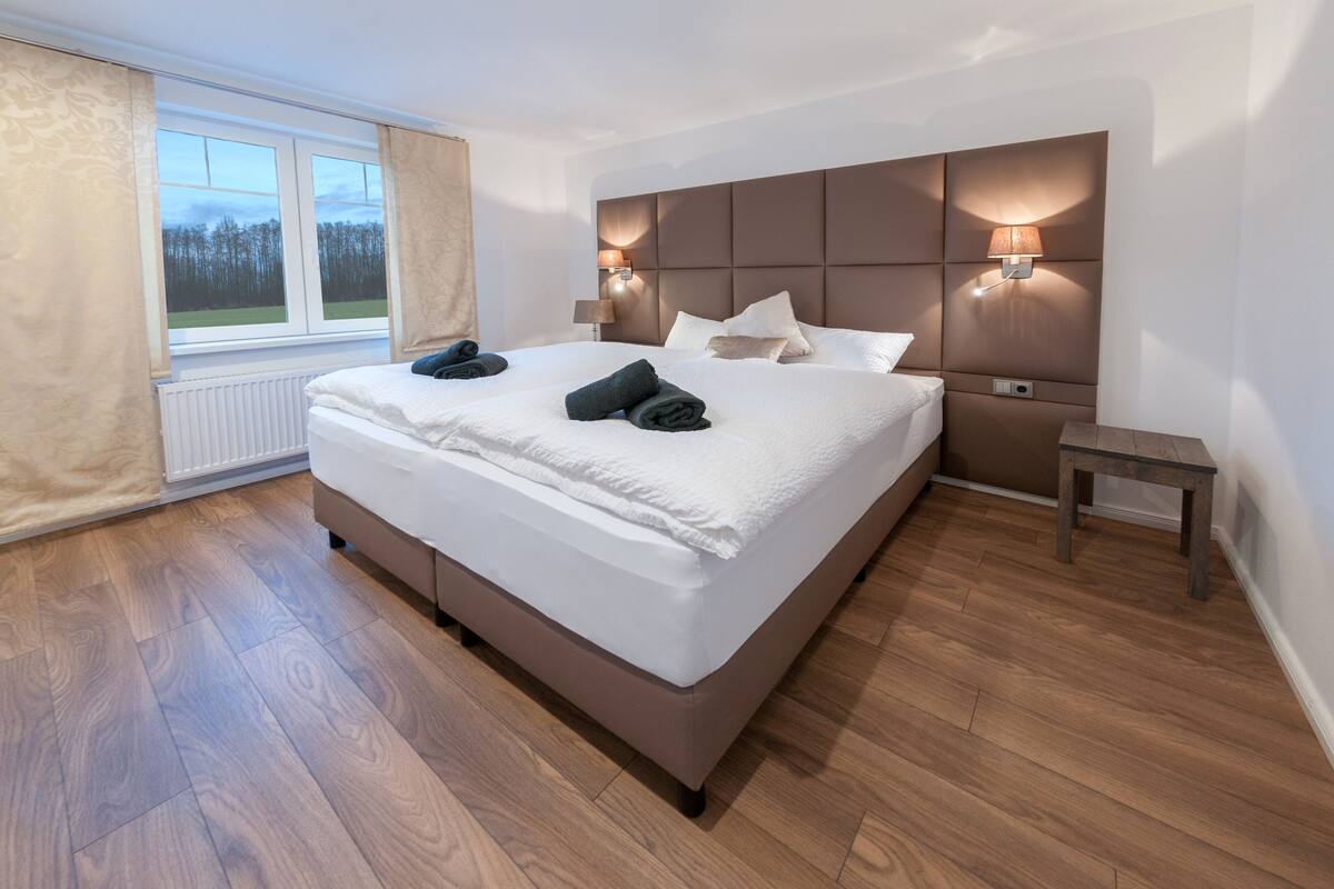 Una habitación de hotel moderna con una cama grande, suelo de madera, una ventana con cortinas e iluminación empotrada.
