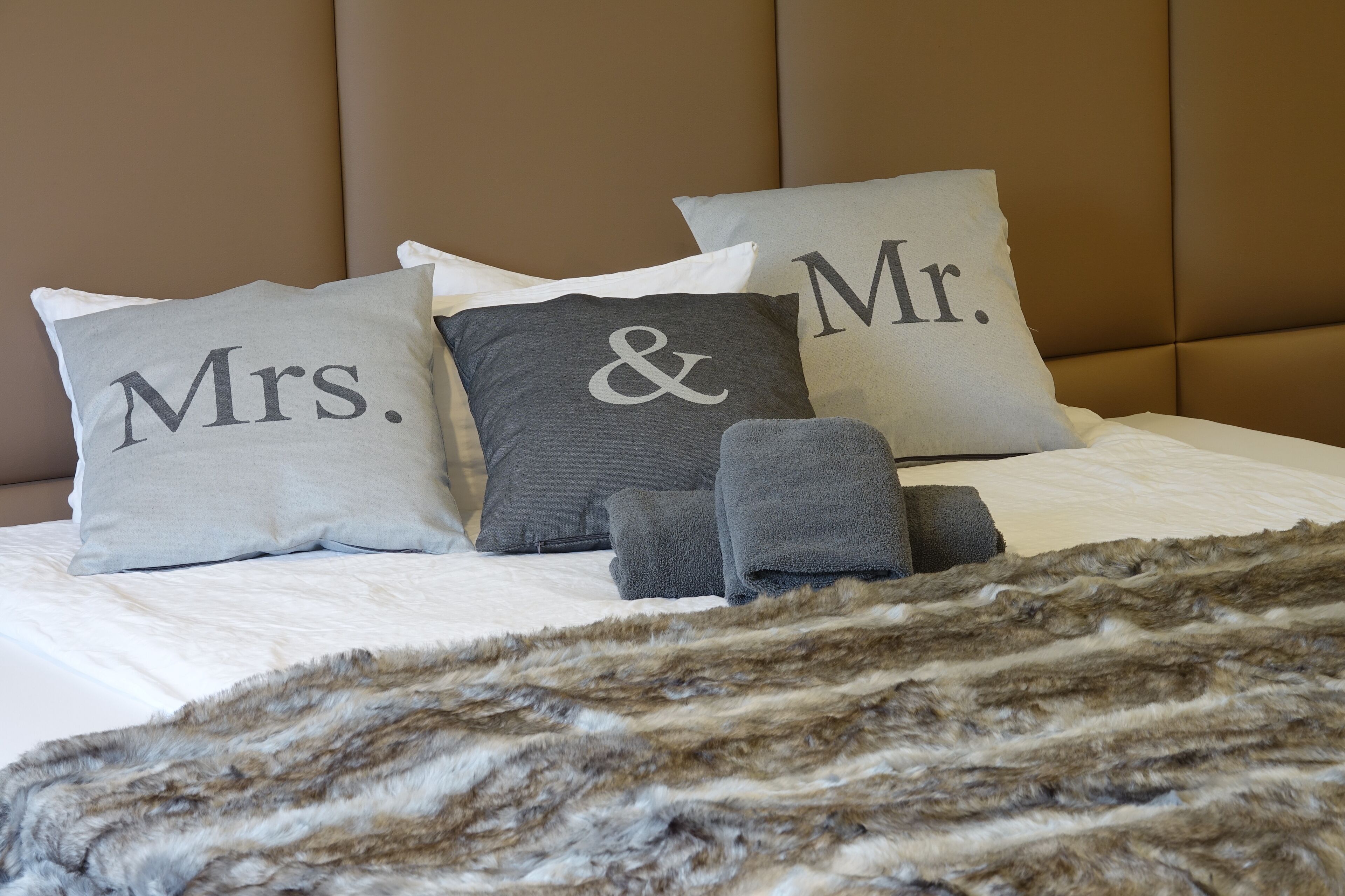 Una cama con tres almohadas etiquetadas como “Mrs.”, “&” y “Mr.”, y un par de toallas grises.
