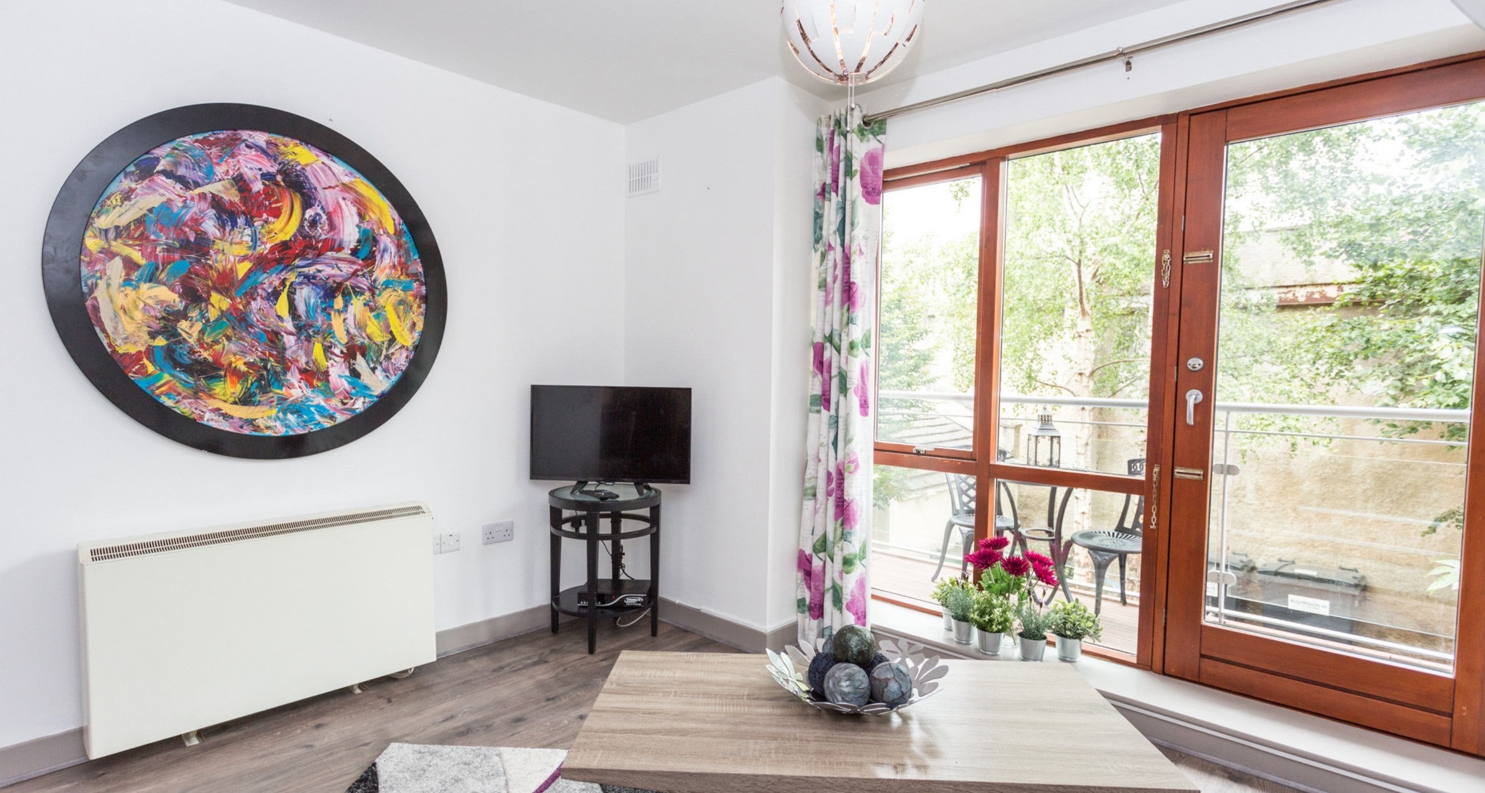 A modern living room with a large abstract painting, a flat-screen TV, a wooden coffee table, and a view of the outdoors through a sliding glass door.