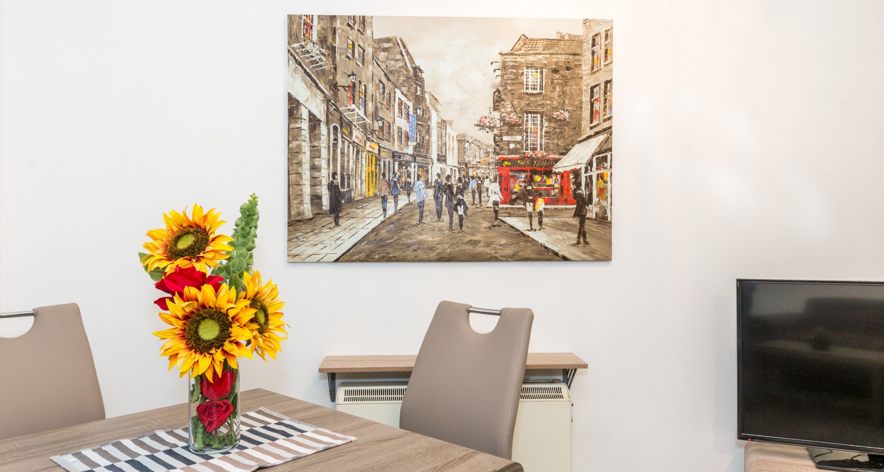 A dining area with a table, chairs, a vase with sunflowers, a painting of a street scene, and a television.