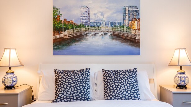 Una habitación de hotel con una cama, dos mesitas de noche, dos lámparas y un cuadro de un paisaje urbano sobre la cama.