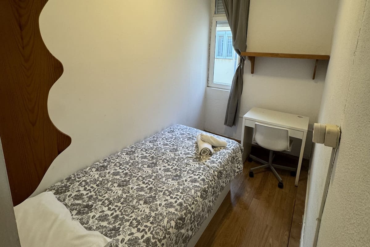 Una habitación pequeña con cama individual, ventilador de techo, escritorio y ventana con cortinas.
