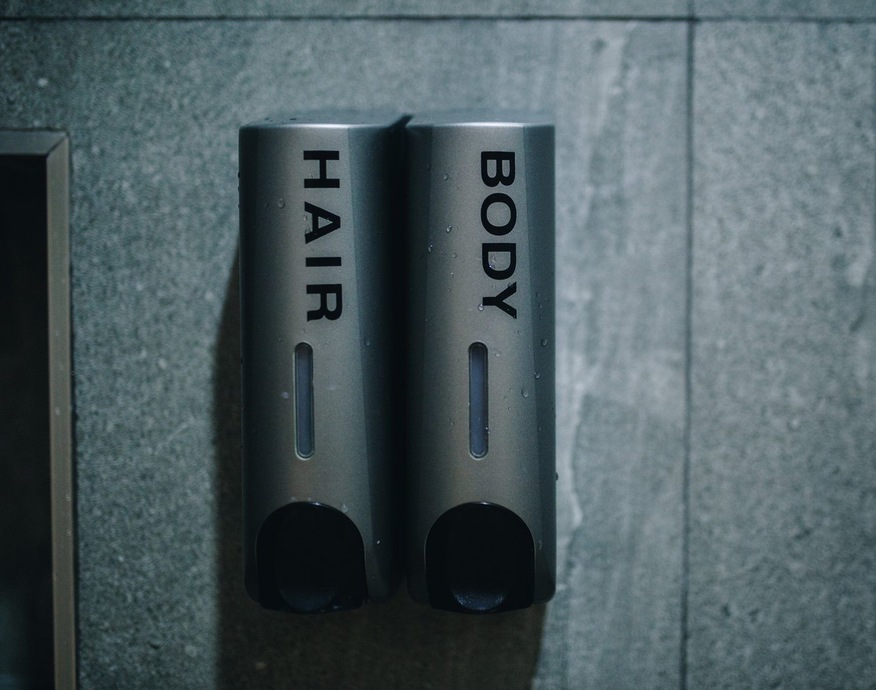 Twee metalen dispensers, aangeduid met 'HAIR' en 'BODY', op een gestructureerde muur.