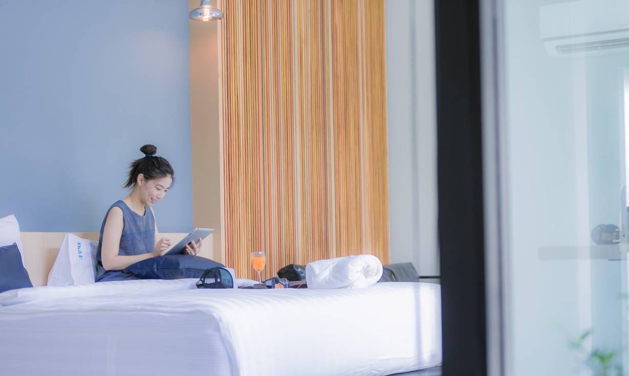 A person sitting on a bed in a hotel room, using a tablet.