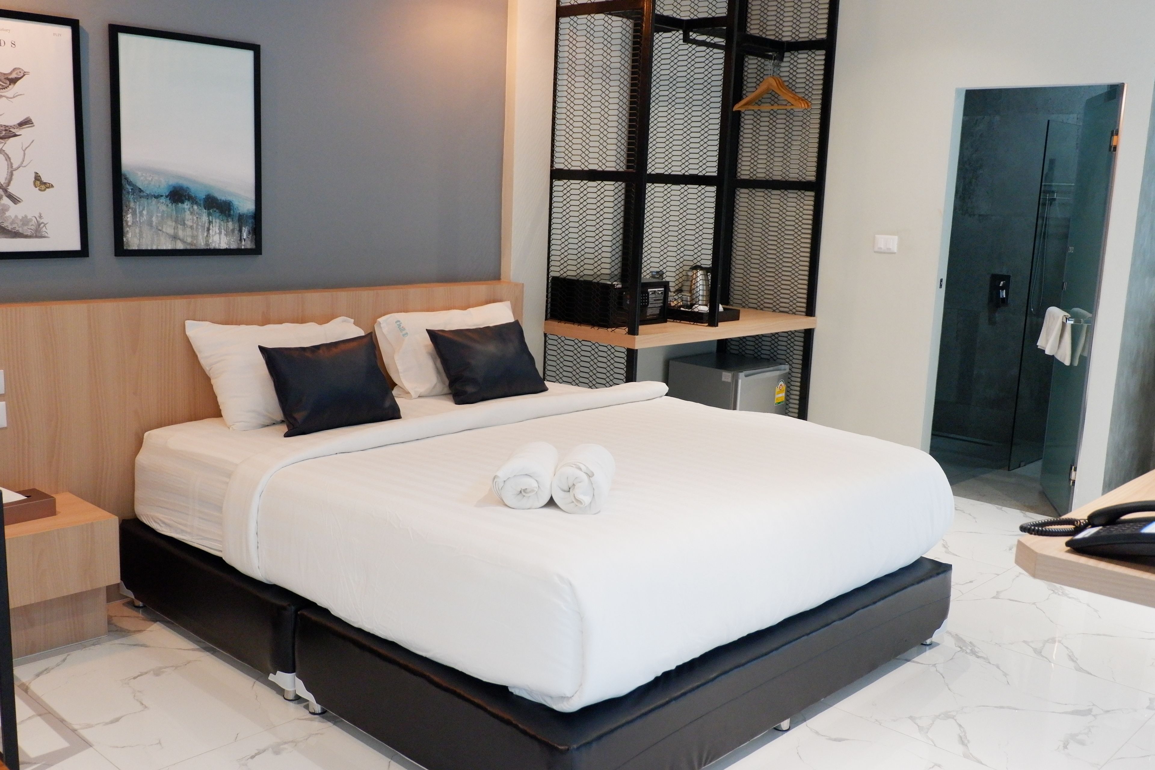 A modern hotel room with a large bed, a wooden headboard, a small bedside table, a built-in shelf, and a glass shower enclosure.