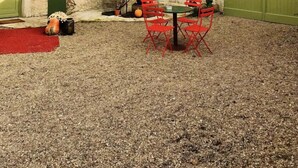 Un patio con una mesa y sillas rojas, rodeado de un muro de piedra y un edificio de ladrillo con contraventanas verdes.