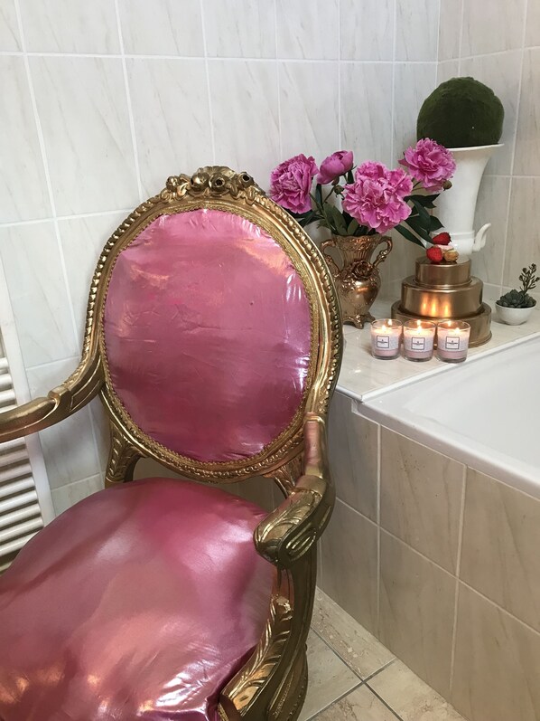 A bathroom with a golden ornate chair, a pink cushioned chair, a vase with pink flowers, and a white bathtub. - Taty Ginette (Marolles)