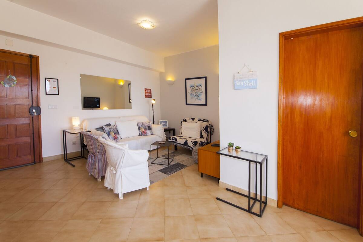 Una sala de estar con un sofá, una mesa de centro y una mesita auxiliar con una lámpara. Se observa una puerta de madera y un cuadro en la pared.