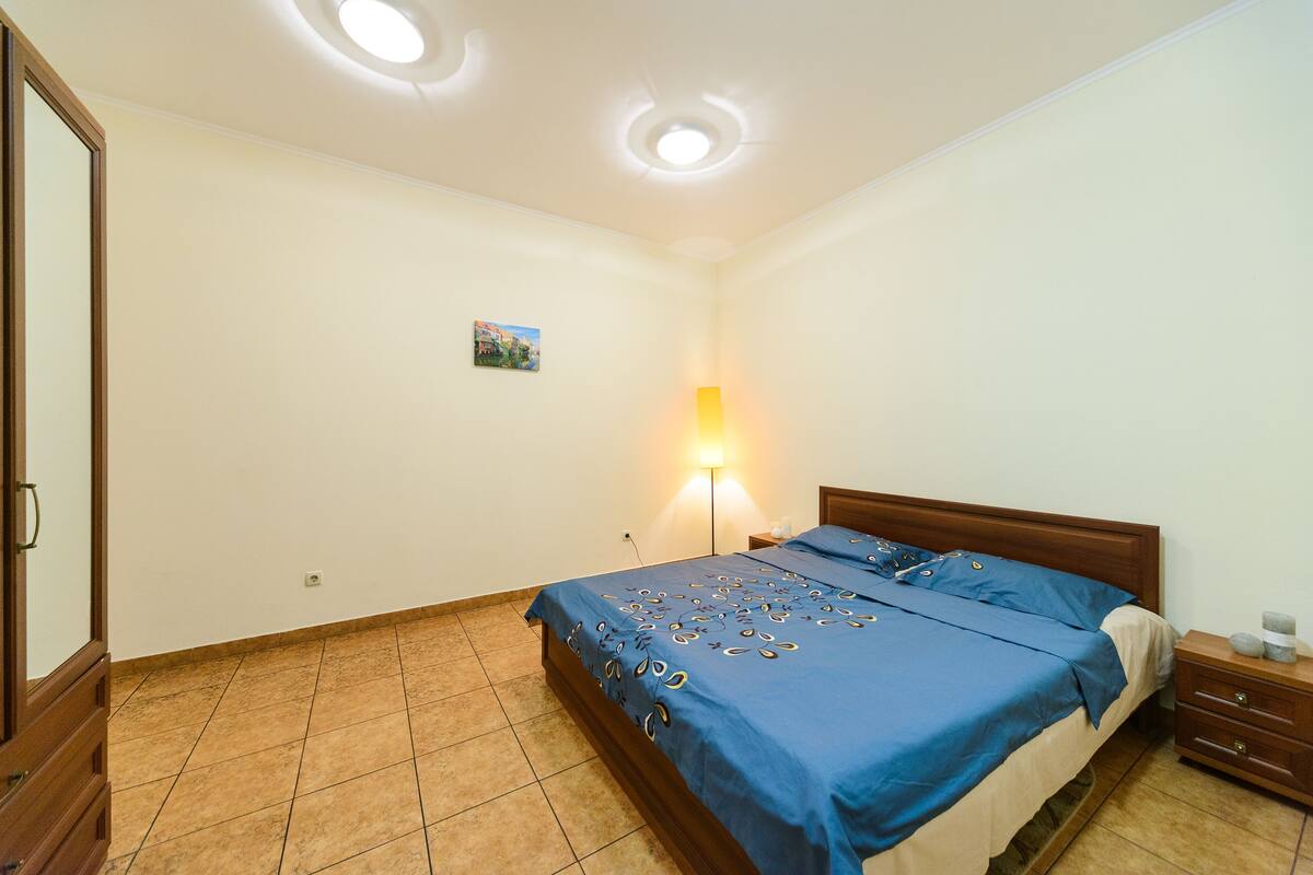 A bedroom with a bed, bedside table, lamp, and a small picture on the wall.