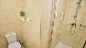 A bathroom with beige tiles, a toilet, a shower with a glass door, and a small shelf with a plant. - The George Hotel (Axminster)